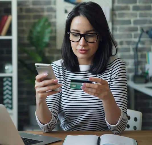 Woman using smartphone and holding a credit card while sitting at a desk with a laptop and notebook.